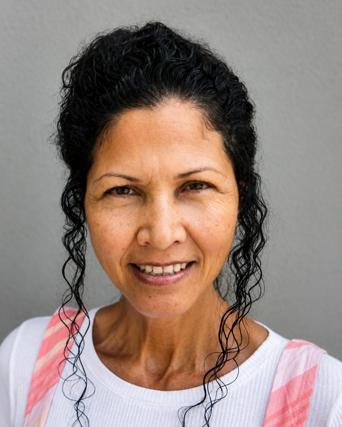 Smiling woman with dark curly hair wearing a white top against a gray background.